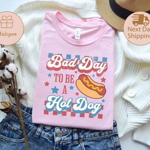 May include: A light pink t-shirt with the text "Bad Day to be a Hot Dog" in a retro font, featuring a hot dog graphic. The shirt is displayed on a white sweater and blue jeans.