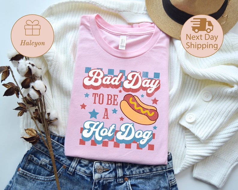 May include: A light pink t-shirt with the text "Bad Day to be a Hot Dog" in a retro font, featuring a hot dog graphic. The shirt is displayed on a white sweater and blue jeans.
