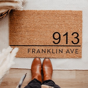 May include: A rectangular coir doormat with the address "913 FRANKLIN AVE" in black lettering. The mat is a natural brown color and is placed on a white rug. Brown leather boots are visible at the bottom of the image.