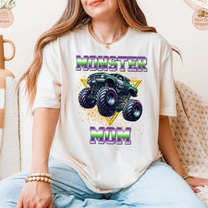 May include: Cream-colored t-shirt featuring a graphic of a green monster truck with purple and black wheels, set against a yellow triangle. The text "MONSTER MOM" is printed above and below the truck in purple and green.