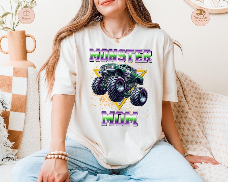 May include: Cream-colored t-shirt featuring a graphic of a green monster truck with purple and black wheels, set against a yellow triangle. The text "MONSTER MOM" is printed above and below the truck in purple and green.