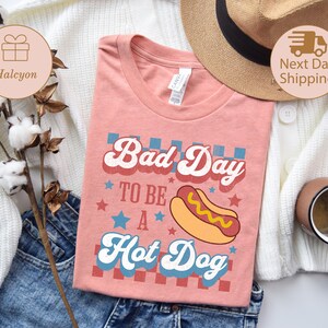 May include: A coral-colored t-shirt with the text "Bad Day to be a Hot Dog" in a retro font, featuring a hot dog graphic. The shirt is folded on a pair of blue jeans, with a straw hat and a coffee mug in the background.