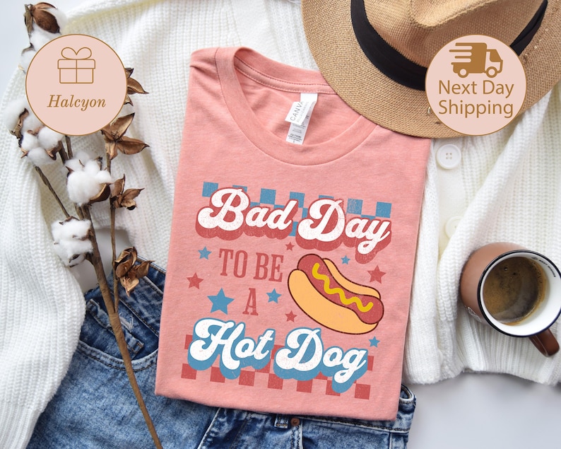 May include: A coral-colored t-shirt with the text "Bad Day to be a Hot Dog" in a retro font, featuring a hot dog graphic. The shirt is folded on a pair of blue jeans, with a straw hat and a coffee mug in the background.