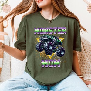 May include: Olive green t-shirt featuring a graphic of a green monster truck with purple and black wheels, and the text "MONSTER MOM" in purple and green. The design includes a yellow triangle and splatter effects.