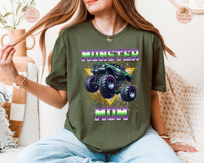 May include: Olive green t-shirt featuring a graphic of a green monster truck with purple and black wheels, and the text "MONSTER MOM" in purple and green. The design includes a yellow triangle and splatter effects.