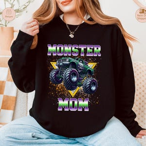 May include: Black sweatshirt with a graphic of a green monster truck with purple and green accents, and the words "MONSTER MOM" in purple and green. The design is set against a yellow triangle.