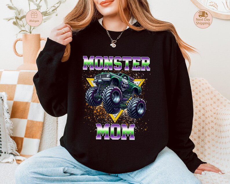 May include: Black sweatshirt with a graphic of a green monster truck with purple and green accents, and the words "MONSTER MOM" in purple and green. The design is set against a yellow triangle.