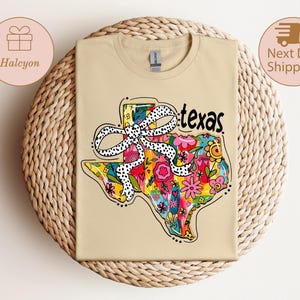 以下が含まれることがあります： テキサス州の形をしたカラフルなデザインが特徴のベージュ色のTシャツ。花柄と水玉模様のリボンが描かれています。「texas」の文字は黒で書かれています。Tシャツは織られた表面に折りたたまれています。