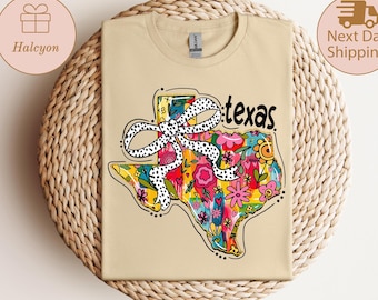 May include: A beige t-shirt with a colourful Texas-shaped design, filled with flowers and a polka-dot bow. The word "texas" is written in black script. The shirt is folded on a woven surface.