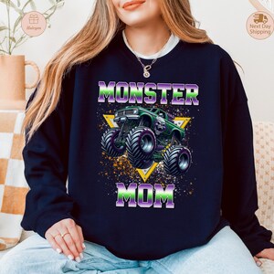 May include: Navy blue sweatshirt with a graphic of a green monster truck and the words "MONSTER MOM" in purple and green. The design is set against a yellow triangle and a speckled background.