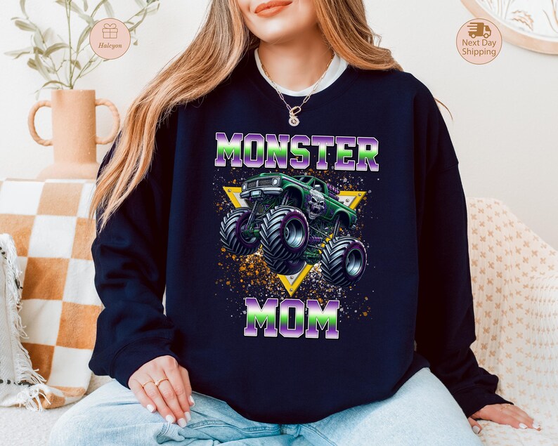 May include: Navy blue sweatshirt with a graphic of a green monster truck and the words "MONSTER MOM" in purple and green. The design is set against a yellow triangle and a speckled background.