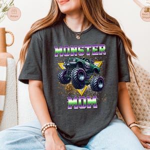 May include: Dark gray t-shirt featuring a graphic of a green monster truck with purple accents, set against a yellow triangle. The text "MONSTER MOM" is displayed in purple and green. The shirt is a casual, crew neck style.