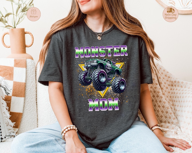 May include: Dark gray t-shirt featuring a graphic of a green monster truck with purple accents, set against a yellow triangle. The text "MONSTER MOM" is displayed in purple and green. The shirt is a casual, crew neck style.