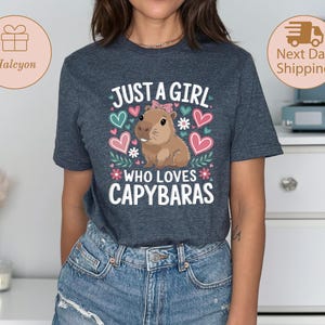 May include: A dark gray t-shirt with the text "JUST A GIRL WHO LOVES CAPYBARAS" in white, pink, and green. The design includes a cartoon capybara, hearts, and floral accents. The shirt is made of a soft material.