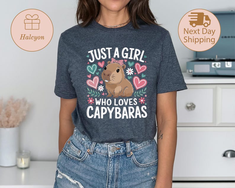 May include: A dark gray t-shirt with the text "JUST A GIRL WHO LOVES CAPYBARAS" in white, pink, and green. The design includes a cartoon capybara, hearts, and floral accents. The shirt is made of a soft material.