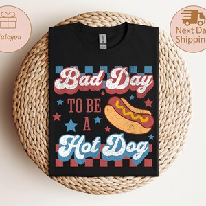 May include: Black t-shirt with the text "Bad Day To Be A Hot Dog" in red, white, and blue retro font. A cartoon hot dog is featured below the text. The shirt is displayed on a woven surface.