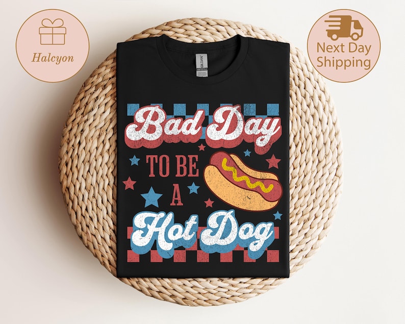 May include: Black t-shirt with the text "Bad Day To Be A Hot Dog" in red, white, and blue retro font. A cartoon hot dog is featured below the text. The shirt is displayed on a woven surface.