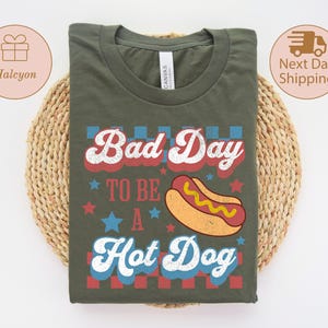 May include: Olive green t-shirt with the text "Bad Day To Be A Hot Dog" in red, white, and blue. The design includes a cartoon hot dog and stars. The shirt is displayed on a woven, circular mat.