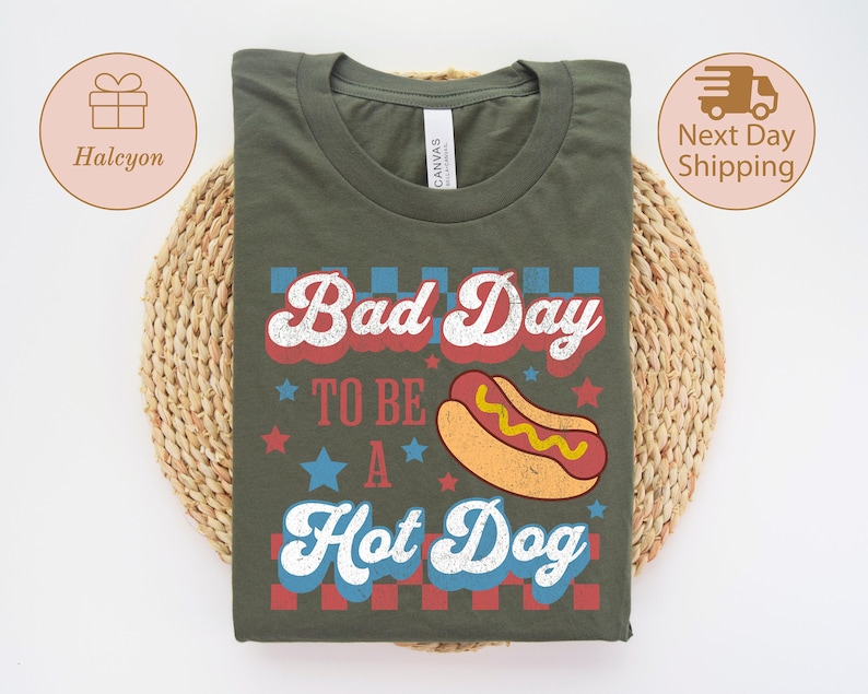 May include: Olive green t-shirt with the text "Bad Day To Be A Hot Dog" in red, white, and blue. The design includes a cartoon hot dog and stars. The shirt is displayed on a woven, circular mat.