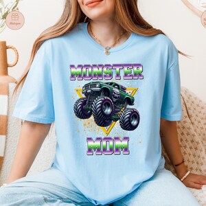 May include: Light blue t-shirt with the words "MONSTER MOM" in purple and green, featuring a green monster truck graphic inside a yellow triangle. The shirt has short sleeves and a crew neck.