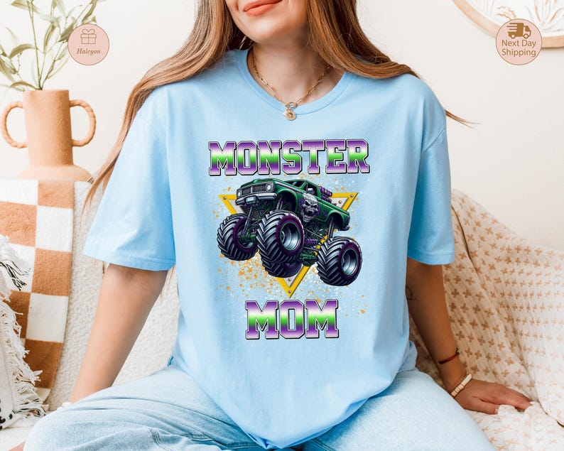 May include: Light blue t-shirt with the words "MONSTER MOM" in purple and green, featuring a green monster truck graphic inside a yellow triangle. The shirt has short sleeves and a crew neck.