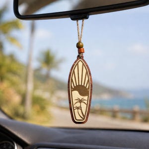 May include: A surfboard-shaped wooden car air freshener hanging from a rearview mirror. The ornament features a laser-engraved design of a sun rising over palm trees, suspended by a jute twine cord with two small wooden beads in brown and tan.