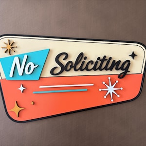 May include: A trapezoidal wall sign with a cream top and orange bottom section. It features the text No Soliciting in black and white script. The design includes turquoise and orange starburst accents, two horizontal lines, and a black border.