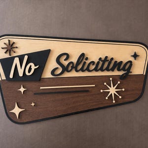 May include: A trapezoidal wooden sign with a black border, featuring the text "No Soliciting" in a layered design. The top section is light wood, the bottom is dark wood, and the sign is decorated with starburst shapes in black and light wood.