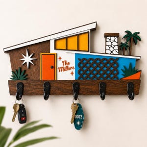 May include: A wall-mounted wooden key holder shaped like a mid-century modern house. It features a dark wood base with five black metal hooks, an orange door, yellow windows, a blue wall with a lattice pattern, and the text The Millers.