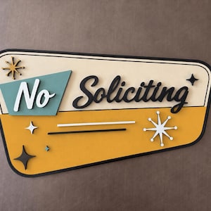 May include: A trapezoidal wall sign with a cream and mustard yellow background featuring the text No Soliciting in black and white script. The sign includes decorative starburst accents in black, white, and teal, all framed by a thick black border.