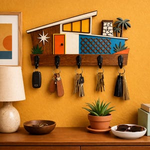 May include: A mid-century modern style key holder in the shape of a house. The wooden key rack features a brown roof, orange door, and blue accents. Black hooks hold keys and keychains. The background is a mustard yellow wall.