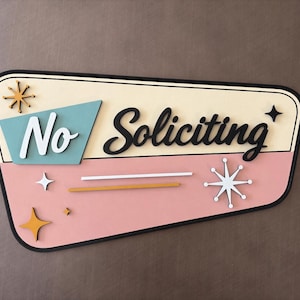 May include: A trapezoidal wall sign with a cream and pink background featuring the text No Soliciting in black and white script. The design includes a teal accent shape, gold and white starburst icons, and two horizontal lines in white and gold.