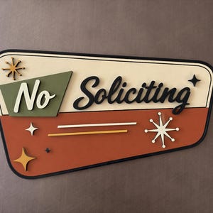 May include: A trapezoidal wall sign with a cream top and terracotta bottom section. It features the text "No Soliciting" in black and white script, accented by olive green geometric shapes and several gold and white starburst icons.