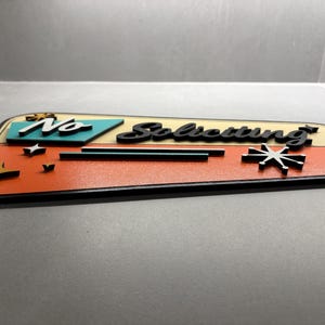 May include: A trapezoidal sign featuring the text "No Soliciting" in black script lettering. The sign has a cream-colored upper section and a textured orange lower section, accented with teal geometric shapes and black starburst icons.