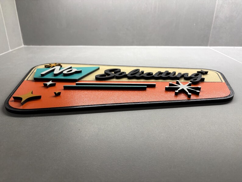 May include: A trapezoidal sign featuring the text "No Soliciting" in black script lettering. The sign has a cream-colored upper section and a textured orange lower section, accented with teal geometric shapes and black starburst icons.