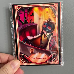 May include: A rectangular trading card in a clear plastic sleeve featuring a character with a television for a head, wearing a vest and tie, sitting at a bar. The card has a red and orange colour palette, a small drink, and the text The Rejection.