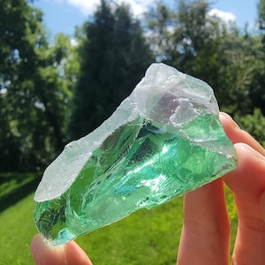 May include: A large, irregularly shaped piece of green glass, held up against a backdrop of green grass and trees under a blue sky. The glass has a translucent quality, with a clear, white edge. The light catches the glass, creating a shimmering effect.