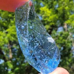 Könnte beinhalten: Ein Stück blaues Glas mit einer rauen, unregelmäßigen Form. Das Glas wird zwischen zwei Fingern gehalten und hat viele kleine Blasen im Inneren. Der Hintergrund ist verschwommenes grünes Laub.