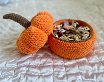 May include: An orange crocheted pumpkin with a brown stem next to a matching bowl filled with wrapped candies. The pumpkin and bowl are on a white, textured surface, suggesting a fall or Halloween theme.