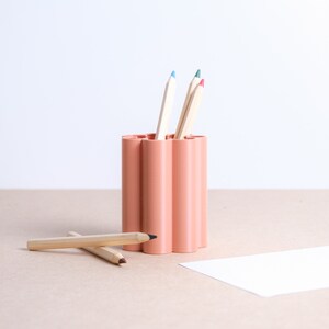 Peut inclure: Un porte-crayons pêche avec un design cannelé unique. Plusieurs crayons de couleur sont à l'intérieur du support, avec quelques crayons posés sur la surface. Les crayons ont des pointes bleues, vertes et rouges. Un morceau de papier blanc est à droite.