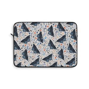 May include: A rectangular laptop sleeve featuring a repeating pattern of dark blue butterflies, crescent moons, and floral elements in orange, blue, and green on a cream background. The sleeve has a black zipper and trim.