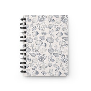 May include: A white spiral notebook with a blue and white seashell pattern. The cover features a variety of seashells, including oysters, clams, and starfish.