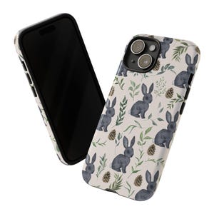 May include: A white phone case with a black border and a pattern of watercolor bunnies, leaves, and pine cones. The case has a cutout for the camera lens.