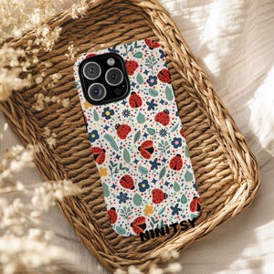 May include: A white phone case with a colorful pattern of red ladybugs, blue and yellow flowers, and green leaves. The case has a black camera lens and the word "NINITSY" printed at the bottom. The case is resting on a woven wicker tray.