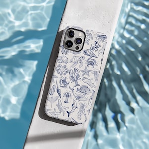 May include: A white iPhone case featuring a blue and white pattern of sea creatures, such as a turtle, starfish, octopus, and fish. The case has a camera lens cutout and a white background.