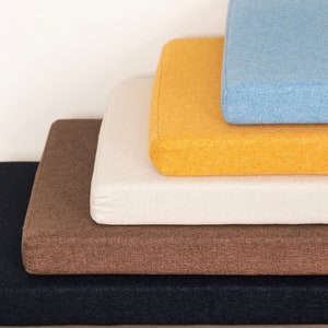 May include: A stack of four cushions in different colors and fabrics. The bottom cushion is black, the next is brown, the third is white, and the top cushion is yellow with a blue top.