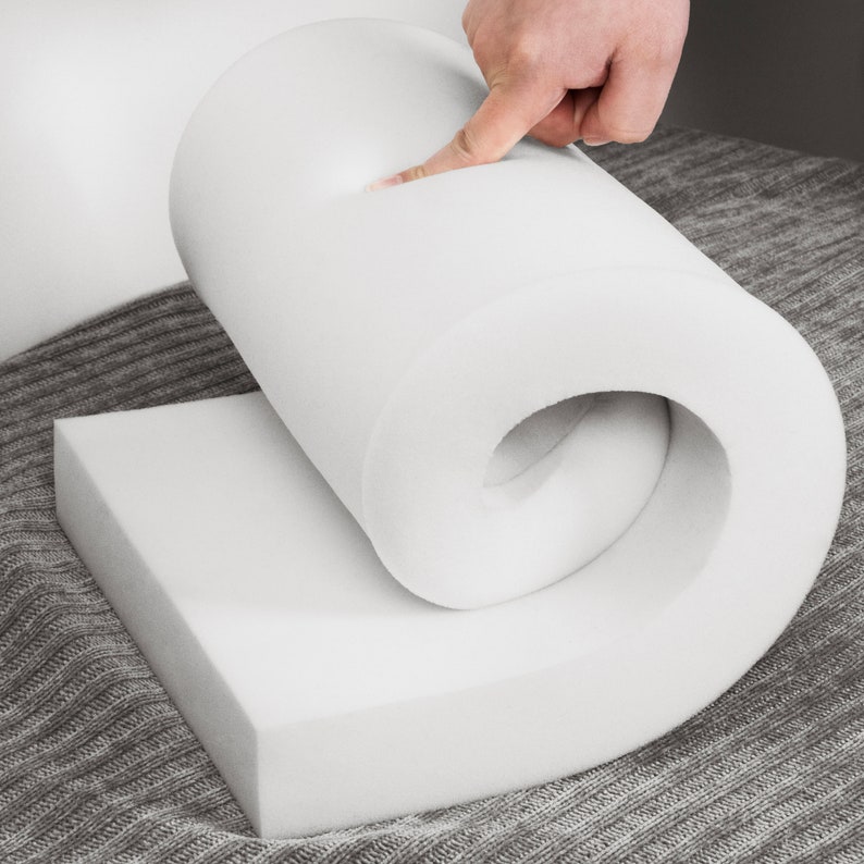 May include: A white foam cushion is rolled up and held by a hand. The foam is dense and appears to be high quality. The cushion is on a gray fabric surface.
