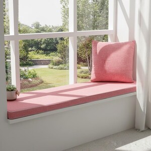May include: A window seat with a pink cushion and matching pillow. The seat is built into a white window frame, with a view of a green yard and trees outside. A small potted succulent sits on the window sill.