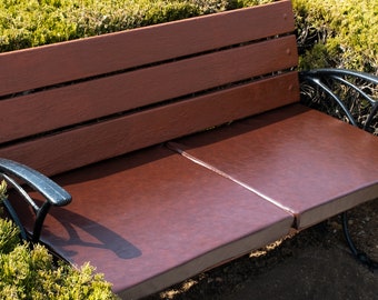 Puede incluir: Un banco de madera marrón con un asiento de cuero marrón oscuro y brazos de metal negro. El banco está sentado en un lecho de hierba verde.
