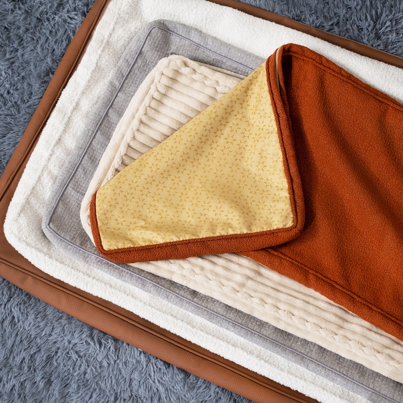 May include: A set of four dog beds, each with a different color and texture. The top bed is a rich orange, the next is a light beige with a ribbed texture, the third is a light gray, and the bottom is a brown faux leather.